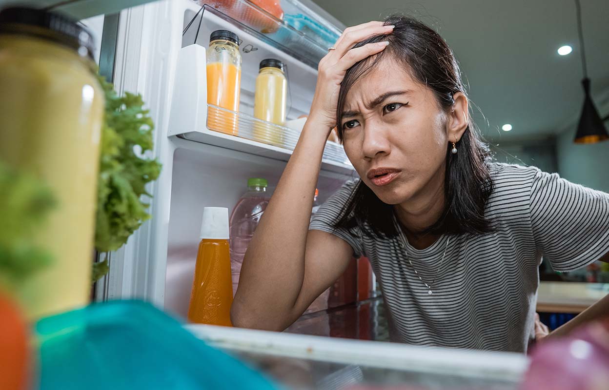 Worried woman looking into an open fridge, noticing food that doesn’t feel cold and suspecting her fridge is failing